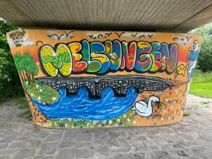 Das fertiggestellte Graffiti auf der dem Weg zugewandten Seite der Brückenstütze setzt einen farbenfrohen Akzent am Fuldaufer. Die Gestaltung ist Teil des KOMPASS-Prozesses zur Aufwertung eines als Angstraum wahrgenommenen Bereichs.