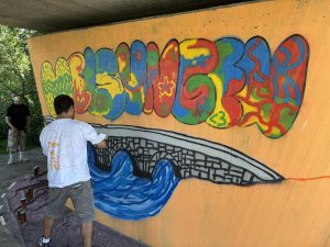 Von der Idee zur Spraydose: Graffiti-Künstler Marcel de Medeiros aus Kassel unterstützt die Schülerinnen und Schüler der Gesamtschule Melsungen mit professionellen Tipps und kreativen Anregungen bei der Gestaltung der Betonstütze unter der Schlossbrücke. Foto: Stadt Melsungen / Alexander Dupont